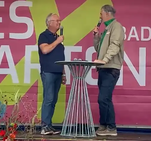 Zwei Männer sprechen mit Mikrofonen auf einer Bühne vor einer bunten Landesgartenschau-Kulisse.